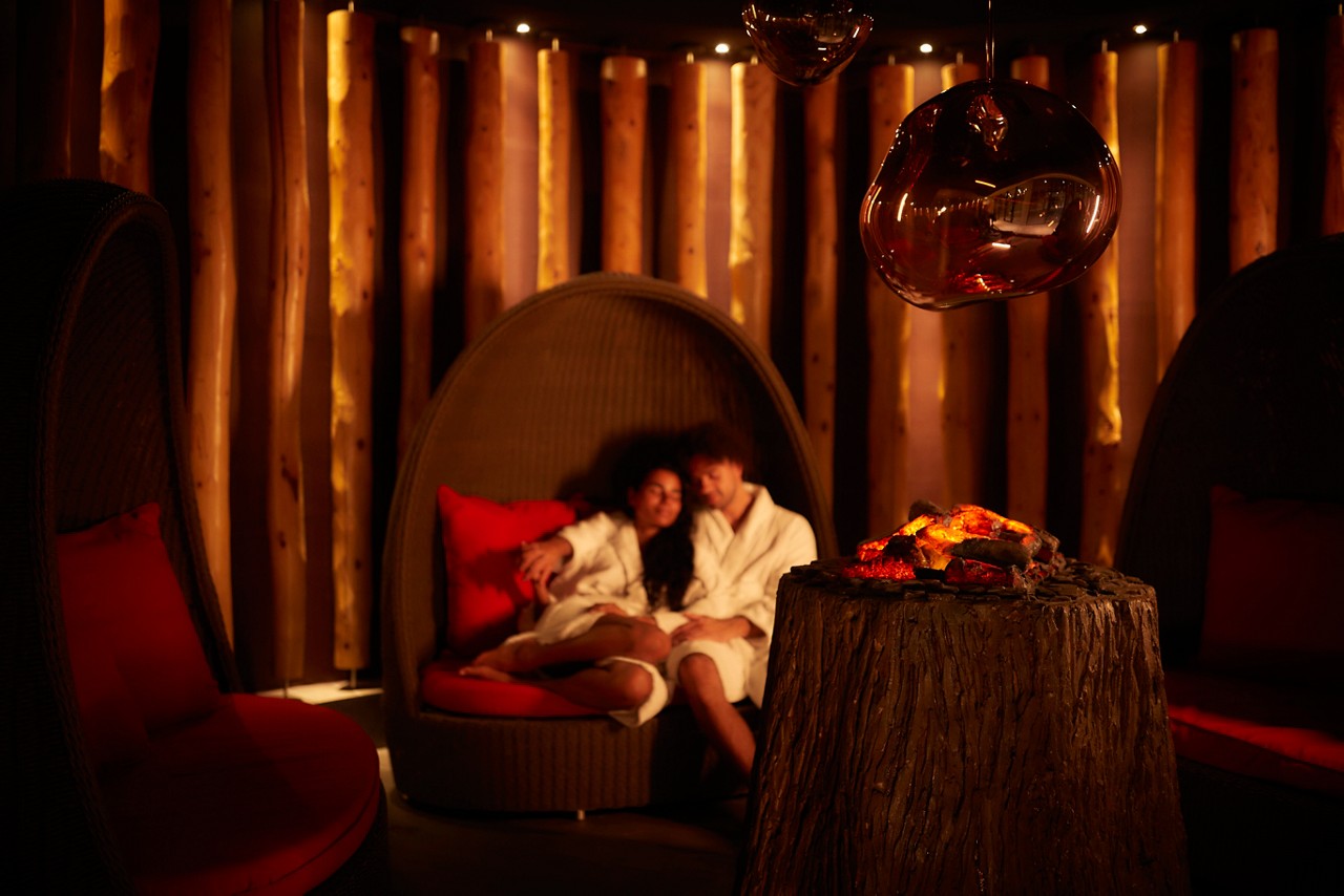 Man and woman snuggled up in a large cocoon chair by a glowing fire.